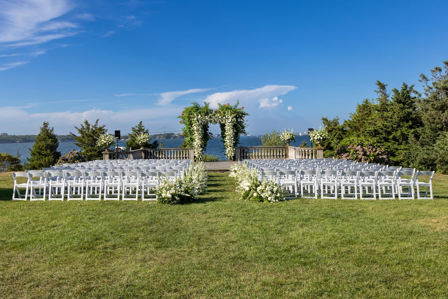 Outdoor waterside wedding ceremony with white floral arch and chairs